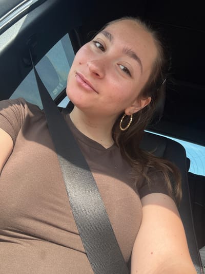 The image shows a woman sitting in the driver's seat of a car, wearing a brown t-shirt and a seatbelt. She is looking at the camera and smiling slightly. The interior of the car is visible, including the dashboard and the side window with a sunshade. The woman has long brown hair and is wearing gold hoop earrings. The lighting suggests it is daytime, with sunlight coming through the window. There are no other people or objects of note in the immediate vicinity. The scene is a typical interior of a vehicle.