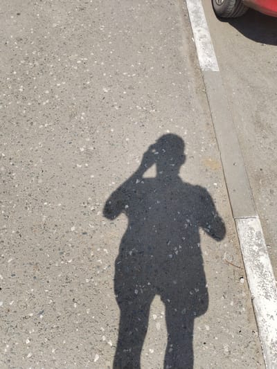 In the city of Troitskoye, Russia, a shadow of a person is cast upon a gray, gravel-speckled asphalt surface. The shadow, elongated and dark, shows the silhouette of an individual holding a phone to their ear. To the right, a concrete curb with white paint markings borders the asphalt. A portion of a red vehicle is visible at the upper right corner of the image, with a tire also present. The scene is illuminated by sunlight, creating a distinct shadow.