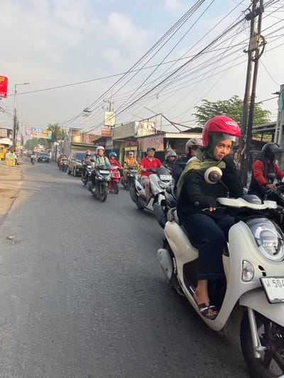 A medium shot captures a street scene in Kedungkendo, Indonesia, with several people on motorcycles in traffic. The perspective is from behind a light-colored scooter, on which a woman wearing a red helmet and a green headscarf is seated, looking to her right. Her left hand is resting on her chin. Behind her, other motorcyclists are visible, some with passengers. The road is paved with asphalt and shows signs of wear. On either side of the street, there are buildings, some with signs indicating businesses, and various utility poles with a complex web of overhead wires. The sky is overcast, suggesting a bright, but possibly hazy, day. The overall impression is one of daily life and transportation in a bustling Indonesian urban or semi-urban environment.