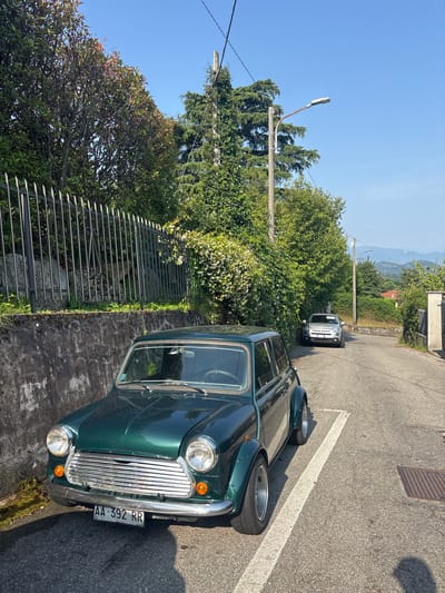 In Orino, Italy, on a clear, sunny day, a dark green classic Mini Cooper is parked parallel to a stone wall along a paved road. The Mini Cooper faces the camera, its chrome grille and round headlights prominent. The road curves gently uphill, with a silver Fiat 500 parked further up on the right side. Lush green trees and foliage line the left side of the road, forming a dense canopy above the Mini. A metal fence runs along the top of the stone wall. In the background, more trees and the gentle slopes of hills under a bright blue sky are visible. The environment suggests a residential or semi-rural area with limited traffic.