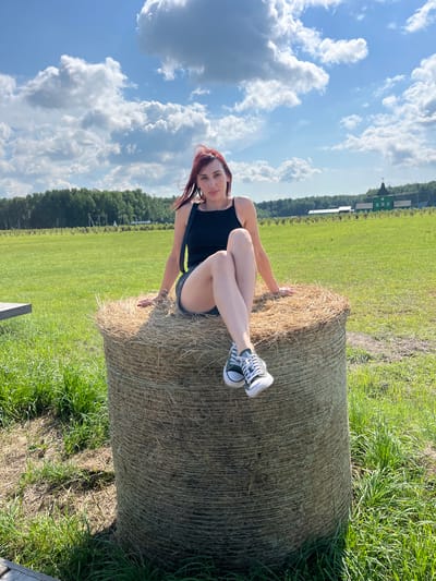 In Akhlebaikha (Kovriginskii S/S), Russia, a young woman with red hair is sitting on a large, round bale of hay in a grassy field under a bright blue sky with fluffy white clouds. She is wearing a black tank top and dark shorts, with black and white Converse sneakers on her feet. Her legs are crossed, and her left hand rests on the hay bale. In the background, a line of green trees stretches across the horizon, and a distinct green building with a pointed roof and weather vane is visible. The scene appears to be a rural setting on a sunny day.