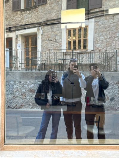 A photograph captures a scene reflected in a window, with the background viewed through the glass confirmed as Fornalutx, Spain. Three individuals are visible in the reflection, each holding a smartphone as if taking a picture or video.
The leftmost individual is a female with long, dark, curly hair, wearing a dark jacket, a white top, and blue jeans. She holds a dark smartphone in both hands, angled towards the viewer. The middle individual is a male with short, dark hair, wearing sunglasses on his head, a dark olive green puffer vest over a white long-sleeved shirt, and dark pants. He also holds a dark smartphone in both hands. The rightmost individual is a male with dark hair, wearing a dark jacket over a white top, holding a dark smartphone in both hands. All three are positioned vertically within the reflection, with the lower half of their bodies visible.
The scene beyond the window features a street or plaza across which stands a multi-story building constructed of light-colored, irregular stone blocks. The ground level of this building includes at least one wooden door and a section with multiple small metal tables and matching chairs, suggesting an outdoor seating area enclosed by a dark metal railing. Above the ground level, upper-story windows are visible, some fitted with dark brown wooden shutters. A prominent yellow rectangular sign is reflected near the top center of the image, appearing above one of these windows. The immediate foreground shows a light-colored wooden window frame partially encompassing the lower left and bottom edges of the image. Visible inside the room from which the photo is taken, reflected in the glass, are indistinct objects, including what appears to be a dark, vertical stand.