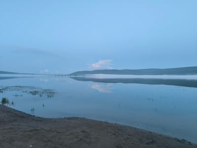 A tranquil lake scene under a pale blue, overcast sky. The water's surface is calm and reflective, mirroring the faint clouds and the distant, low-lying hills that are partially obscured by a thin layer of mist. The foreground features a sandy and pebbly shoreline with some sparse vegetation growing at the water's edge. The overall atmosphere is serene and peaceful, suggesting either early morning or late evening twilight. The image does not contain any people or visible structures that would pinpoint a specific location within the city of Гремучий, Russia.