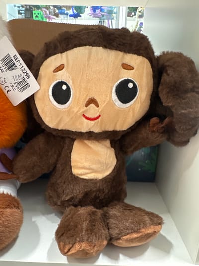 This is a close-up, eye-level, slightly tilted shot of a brown plush monkey toy. The monkey is lying on its back, facing the viewer, with its head slightly to the left. Its face is the most prominent feature, with large, black and white eyes, a tan muzzle, and a small red line representing a mouth. The monkey has fuzzy brown fur and lighter tan patches on its cheeks.

Visible on the lower left of the image is a white tag attached to the monkey. The tag has black text and a barcode. Visible text includes "REF:112298", "1.00", "2980", ".R.C", and "CE".

The monkey is situated within a white shelving unit. In the background, out of focus, there are glimpses of colorful items, possibly toys or decorative objects, on a shelf below. The lighting suggests an indoor setting, likely a store or a display. There are no people visible in the image, and the time of day and weather are not discernible. The overall mood is playful and childlike due to the nature of the subject.