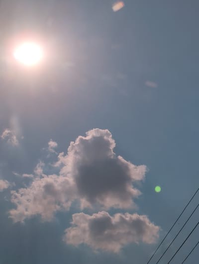 The image depicts a bright, hazy sky with a prominent sun on the left side, casting a warm glow that suggests it is either sunrise or sunset. In the mid-right, a cluster of fluffy, light-colored clouds is visible against the pale blue sky. Above these clouds, a small, vibrant green orb is present, possibly a lens flare or an atmospheric phenomenon. Towards the top right corner, thin, dark, parallel lines stretch across the frame, which appear to be power lines. There is a very faint, dark speck visible near the power lines that could be a bird. The overall impression is one of a serene, natural setting, with the sun and clouds dominating the visual field. There is no visible text in the image.