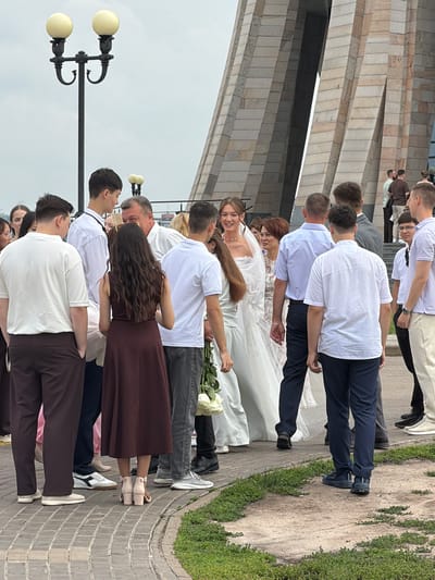 The image captures a wedding party assembled outdoors, possibly at a landmark structure in Vakhitovo, Russia. In the foreground, a group of people are gathered, facing a bride in a white wedding dress and veil, who is smiling. She is holding a bouquet of white roses. Several men in formal attire, including suits and collared shirts, are present. Some women are also in the group, one wearing a dark maroon dress. Behind the wedding party, a large, angular building with a stone facade dominates the background. A lamppost with two globes is visible to the left. The ground is paved with cobblestones and bordered by a patch of green grass. The lighting suggests it is daytime, with an overcast sky.
