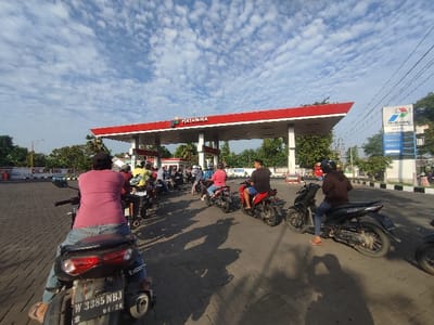 The image shows a gas station in the city of Kecamatan Beji, Indonesia, located at coordinates -7.607, 112.769. The scene is a gas station with a red canopy and a sign that reads "Pertamina." Several motorbikes are parked in a line. People are standing around, some are filling their motorbikes with gas and others are waiting. The environment is paved with a gray concrete block. It is a sunny day with a few clouds in the sky.