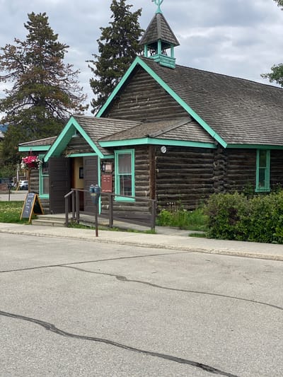 The image shows a rustic log building with a turquoise-painted roof trim and windows, possibly a historical museum, located in Whitehorse, Canada. The building has a steep gabled roof covered with wooden shingles and a small belfry with a cross on top. Double dark brown doors stand open at the entrance, flanked by windows. A ramp leads up to the entrance. To the left of the entrance, a black A-frame chalkboard sign stands on the sidewalk, displaying "WESTERN Yukon MUSEUM OPEN 10-5 DAILY" and "FREE ADMISSION". Hanging baskets with pink and white flowers are suspended above the entrance.
In front of the building, a paved sidewalk runs alongside the street. A wooden railing lines a ramp and stairs leading to the entrance. Two grey parking meters stand on the sidewalk near the entrance. A variety of green bushes and some flowering plants are visible around the base of the building. Two tall evergreen trees are behind the building. The sky is overcast with grey clouds. The street in the foreground is paved with asphalt and shows some cracks and repairs. A white car is parked on the left side of the street in the background.