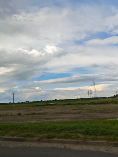 This is a medium shot of a rural landscape under a cloudy sky in the vicinity of Krasnenskoye village council, Belarus. In the foreground, a paved road with a curb runs horizontally across the bottom of the frame, partially blurred by motion. Adjacent to the road is a strip of vibrant green grass, which transitions into a brown, tilled field. In the middle ground, the field stretches towards a distant line of trees and a few buildings, some of which appear to be houses. Several power lines are visible, supported by tall, slender poles, with two larger transmission towers in the right half of the frame. The sky above is filled with a mix of light and dark grey clouds interspersed with patches of blue sky. A faint, almost imperceptible rainbow can be seen in the distance on the left side of the image. The scene is peaceful and conveys a sense of open space.