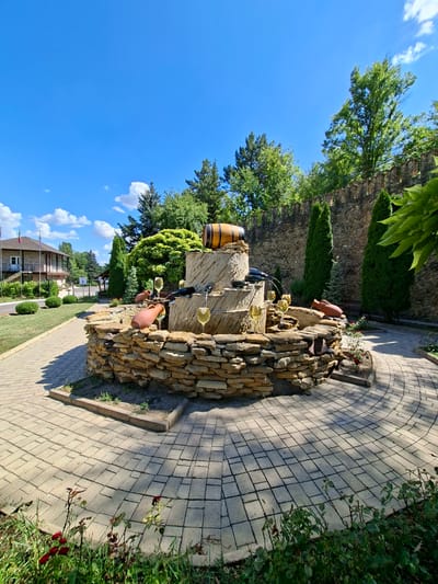 This image captures a sunny day scene outside a building in Milestii Mici, Moldova. A decorative fountain, made of stacked stone and featuring a wooden barrel on top, is the central element. Several wine glasses and clay amphorae are incorporated into the fountain's design, suggesting a theme related to winemaking. Water flows from the fountain, creating a pleasant ambiance.

In the background, a stone wall runs along the property line, with trees and manicured bushes adding to the landscape. A building with a beige facade and balconies is visible on the left, with flags displayed in front. The ground is paved with light-colored bricks, forming a circular pattern around the fountain. Lush green grass and some red roses border the paved area in the foreground. The sky is a clear blue with a few scattered white clouds. No people are visible in the frame.