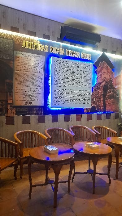 This image depicts an indoor cultural display and seating area, located in Glantengan, Indonesia. No individuals are present. The background wall features a prominent exhibit dedicated to "AKULTURASI BUDAYA MENARA KUDUS" (Cultural Acculturation of Kudus Minaret), comprising two large text panels and a large photographic print of the Kudus Minaret, all accentuated by blue LED backlighting. An air conditioning unit is mounted high on the wall above the exhibit. In the foreground, four dark wooden, round pedestal tables are arranged with matching wooden chairs. Each visible table holds a stack of small, white-covered items resembling books or brochures. The floor is covered with reddish-brown tiles, reflecting the blue LED illumination.
