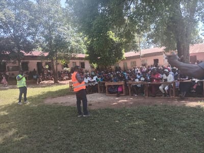An outdoor daytime scene depicts a public gathering in Idakho South ward, Kenya. In the foreground, a grassy area transitions into a dirt path. Two men are standing; one on the left wears a yellow-green reflective vest and dark pants, while another in the center wears an orange reflective vest, a patterned shirt, and dark pants, appearing to address the assembly with his hand raised near his face. A large crowd, estimated to be over 100 individuals, primarily adults, is seated on multiple rows of rustic wooden benches facing the two standing men. Several individuals within the seated crowd are observed clapping. In the background, beneath mature trees, single-story buildings with light-colored walls and reddish-brown roofs are visible, with additional people gathered in front of them. The environment is illuminated by bright sunlight, casting distinct shadows.
