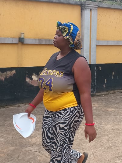 An adult female is depicted in Itu, Nigeria, walking in a side profile view, facing right. She wears a blue, yellow, and black patterned head wrap. Her attire consists of a sleeveless v-neck top that is black at the shoulders, transitions to gray, and becomes yellow at the lower torso, featuring "LAKERS" in yellow text and the number "24" in purple text on the chest. She wears loose-fitting pants with a black and white zebra print and dark-colored sandals. Two red bracelets adorn her left wrist. In her left hand, she holds a white, irregularly shaped plate featuring two red chili pepper designs. The background consists of a solid light orange/dark yellow painted wall with visible gray concrete structural pillars and a small, dark-colored light fixture mounted on the upper left section. The lower portion of the wall shows dark discoloration and some paint deterioration. The ground is a light gray, paved surface.