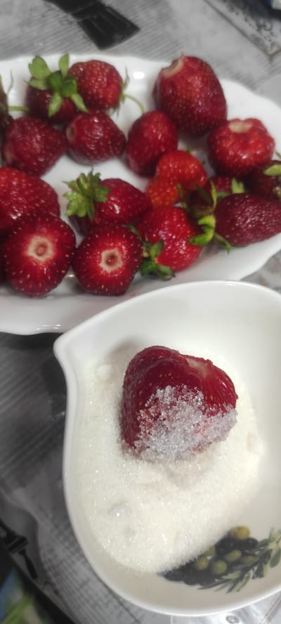 A still life photograph displays a white plate filled with fresh strawberries. In the foreground, a white bowl contains sugar, with a single strawberry partially submerged in it, its bottom half coated in sugar crystals. The scene is set on a surface that appears to be a tablecloth with a subtle checkered pattern. The image is likely captured in Kobeliachok, Ukraine.