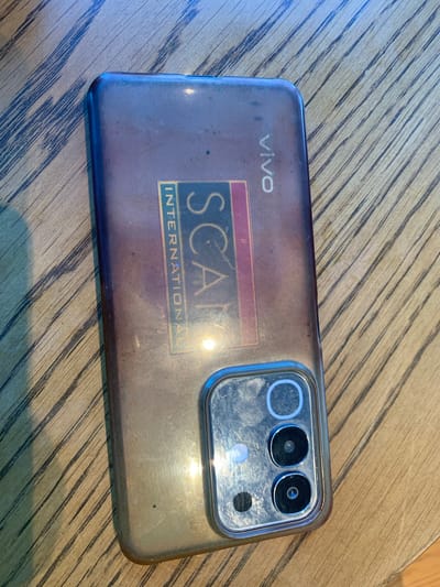 The image shows the back of a smartphone with a distinctive "SCAM INTERNATIONAL" sticker partially covering the camera array and a "VIVO" logo near the top. The phone is placed on a wooden surface with a prominent grain pattern. The lighting suggests it is daytime, casting shadows and highlights on the phone and the table. No specific location in Angeles City, Philippines, can be determined from the image alone.