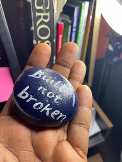 A dark-skinned hand holds a painted rock. The rock is a deep blue color and has the words "Built not broken" written on it in white paint. The background is cluttered with books and other objects on a shelf. A bright light shines on the scene from above, illuminating the hand and the rock. The focus is primarily on the painted rock.