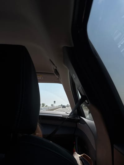 The image captures a forensic perspective from inside a moving vehicle, looking forward and slightly to the right, on a multi-lane highway near Malo Konare, Bulgaria, under a clear, bright daytime sky. The dark-colored rear of a front passenger seat dominates the left foreground, partially obscuring a person with light brown hair. To the right, the vehicle's interior trim, A-pillar, and a black seatbelt are visible. An upward-folded sun visor on the passenger side displays an "AIRBAG" warning label. On the floor or door pocket on the right, a light-colored sandal is present. Through the windshield, an asphalt highway stretches into the distance, populated by other vehicles. A metal guardrail lines the right side of the road, behind which a circular road sign and sparse vegetation are discernible.