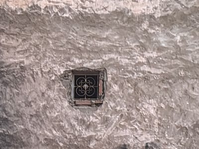 A small, square window with a decorative wrought iron grate is set into a rough, textured, light-colored stone wall. The window is recessed and has a wooden frame that appears aged. The wall surface is uneven and shows signs of wear and age, with various shadows and highlights indicating an irregular texture. The overall impression is one of an old building in a historic setting, possibly in the city of Berrechid, Morocco, given the architectural style.