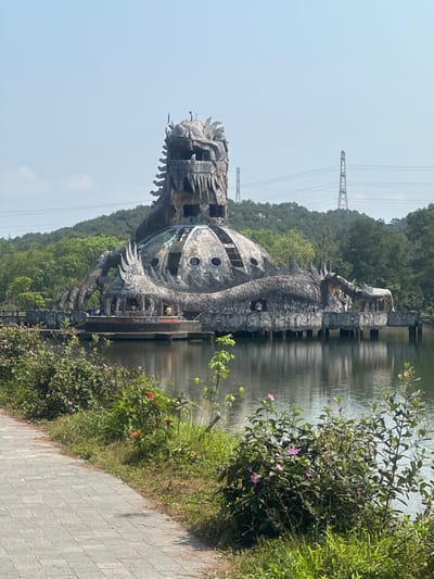The image depicts the abandoned Hồ Thủy Tiên Water Park in Thủy Xuân Ward, Huế, Vietnam. The central subject is a derelict, dragon-shaped concrete structure rising from a calm body of water, likely Thuy Tien Lake.

The primary object is a multi-tiered, weathered concrete edifice representing a large dragon. Its head forms the uppermost section, featuring an open mouth and eroded, tooth-like projections. The body of the dragon forms a dome-like structure below the head, with numerous circular and rectangular openings, indicating former windows or access points. The concrete surfaces exhibit significant discoloration, staining, and material degradation, consistent with prolonged exposure and neglect. The dragon's tail and extremities wrap around the base, which rests on multiple pilings within the water, creating a partially submerged platform. A sign displaying "TRÀ MÈO" (Tea Cat) is visible on a lower, accessible section of the structure.

Several individuals are present on the platforms surrounding the base of the dragon structure, appearing to move about and observe the site. Their exact actions are not distinct due to distance.

The immediate foreground features a paved pathway, composed of light-colored pavers, curving from the lower left to the center. Adjacent to the path is a strip of unkempt grass and various shrubbery, some bearing small pink and orange flowers. The water surface reflects the structure and the clear, light blue sky above.

In the mid-ground and background, dense green foliage covers hillsides, indicating a natural, forested environment. Two metallic lattice transmission towers with visible power lines are present on the distant horizon, rising above the treeline. The scene is illuminated by direct sunlight, casting subtle shadows.