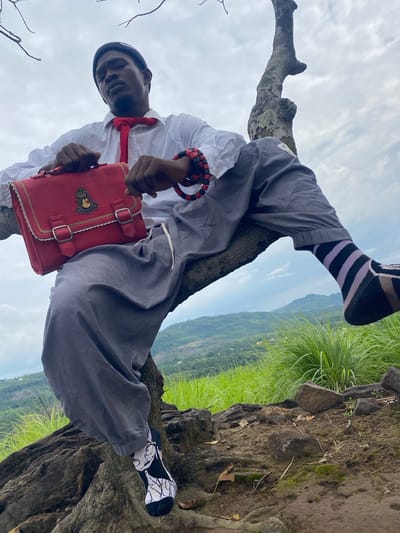 A person is sitting on a tree branch in a natural environment. The person is wearing a white shirt, a red tie, grey pants, and black and white patterned socks. They are holding a red bag with a crest on it. The background shows a landscape with green hills and a cloudy sky. The location is Asaga Amangwu, Nigeria.