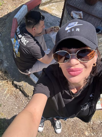 The image, taken from an elevated, near-vertical perspective, depicts two individuals in an outdoor setting in what can be identified as Probuda, Bulgaria.
A person wearing a black Nike Air cap and round sunglasses is positioned towards the top of the frame. They are wearing a black hoodie with white script and a gold necklace, and lipstick, and appear to be holding the camera.
To the left, a second person is seated on a concrete curb with a red and white painted edge. They are wearing a black t-shirt with cartoon characters and the phrase "WHAT'S UP DOC!", light-colored pants, and have sunglasses perched on their head. They are eating food that is wrapped in a plastic bag.
The ground consists of gravel and dry vegetation. In the background, there is a weathered wooden surface with a plastic bag containing remnants of food. The shoes worn by the camera holder appear to be Chuck Taylors. The scene appears to be taken during daylight.