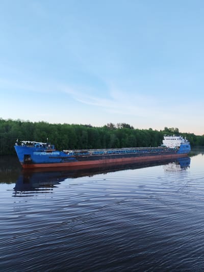 The image captures a large cargo ship, likely a river barge, navigating a calm river at Volokov Most, Russia. The vessel, oriented left to right, features a prominent dark blue hull with a reddish-brown section below the waterline. A white superstructure is visible at the stern. On the bow, the name "СЕВАСТОПОЛЬ" (SEVASTOPOL) is clearly inscribed in white Cyrillic letters.
The river's surface, a deep blue-grey, displays gentle ripples and reflects the ship and the sky. A dense, verdant line of trees forms the backdrop along the far bank, indicating a lush, forested environment. The sky above is a soft, clear light blue with a slight gradient, suggesting either early morning or late afternoon, contributing to the tranquil atmosphere. The weather appears serene and calm as the ship glides through the peaceful waterway.
