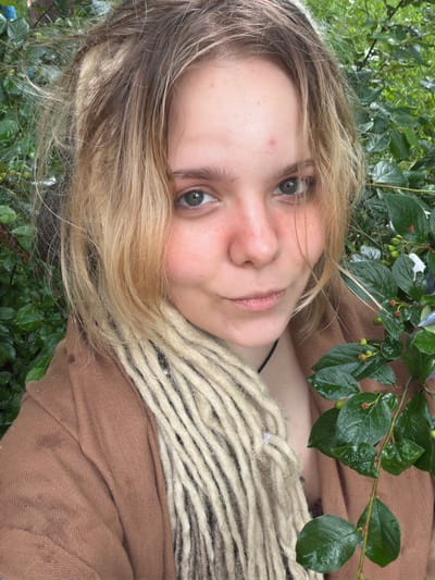 In the Vostochny District of Russia, a young woman with blonde hair styled in dreadlocks is captured in a close-up portrait. She wears a brown jacket and is positioned outdoors, surrounded by lush green foliage. The natural light illuminates her face, highlighting her features and the texture of her hair. The background is filled with the dense green leaves of plants, suggesting a natural or garden setting within the urban landscape.