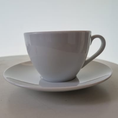 A close-up, low-angle shot focuses on a white ceramic teacup and saucer, presented on a textured, off-white surface that may be a table. The image is rotated, with the cup and saucer appearing on their sides. The cup's handle is prominently featured, arching upwards, and its smooth, glossy surface reflects the ambient light. The saucer is slightly visible beneath the cup, also white and curved. The background is a plain, bright white, suggesting an indoor setting with ample, diffused lighting. There are no people visible in the frame. The overall impression is one of simplicity and cleanliness, highlighting the form and material of the tableware. The lighting creates subtle shadows and highlights, emphasizing the curvature of the cup and saucer. The texture of the surface beneath suggests a matte finish, contrasting with the gloss of the ceramics. There is no visible text in the image. The time of day, weather, and specific location within Jerez are not discernible from the visual information provided.