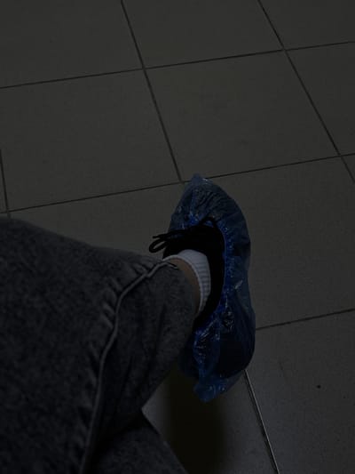 The image shows a person's left leg, clad in dark gray jeans, with a white sock and a dark-colored shoe covered in a blue plastic shoe cover. The person is seated on a floor tiled with dark gray square tiles. The scene appears to be indoors, possibly in a medical facility or a clean room environment, given the shoe cover. There is no indication of the specific location within the city of Rezh, Russia. The lighting is dim, casting shadows on the tiled floor.