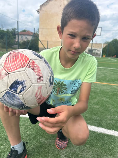 A young boy sits on a green artificial turf field, likely a soccer field, holding a soccer ball. He is wearing a light green t-shirt with a tropical-themed design and dark shorts. He has dark hair and a fair complexion. The soccer ball is predominantly white with blue and red patterned panels. The boy is looking directly at the camera with a slight smile. In the background, a building and trees are visible. The location may be in the city of Kalchevo, Bulgaria.