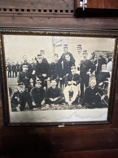 A black and white, framed photograph displays a group of approximately 20 men in uniform, likely military or police, standing and sitting in an outdoor setting with grass and what appears to be a dirt or gravel ground. The men are wearing dark-colored jackets with high collars, matching hats, and some have light-colored pants or breeches. Many of the men have mustaches. Some are holding staffs or rifles. In the background, there are hints of structures, possibly tents or buildings, and more individuals are visible further back. The photograph shows signs of age, with some staining and fading on the image itself. The frame is ornate, with dark wood and gold detailing. The photograph is displayed on a wooden surface, likely a table or shelf, and a portion of a wooden wall or cabinet is visible above it. The context of Mesjidpirak, Indonesia, is not directly inferable from the visual elements within the photograph itself.