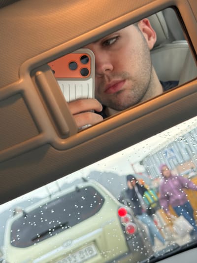 A man takes a mirror selfie in a car. The reflection also captures a view of a street in Ondara, Spain. Raindrops are on the glass. In the street, there is a pedestrian wearing a purple jacket and another one with a colorful shirt. A vehicle is parked nearby.