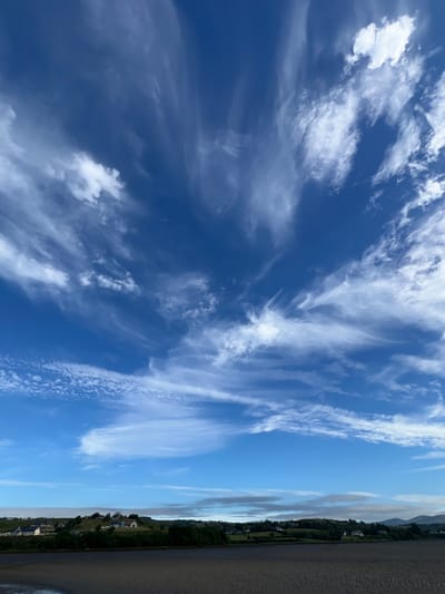 The image depicts a wide expanse of a blue sky filled with wispy, white clouds. Below the sky is a green, rolling landscape dotted with several white houses. In the foreground, a dark, sandy beach stretches across the bottom of the frame. The scene is tranquil and appears to be a coastal view in Tullycullion, Ireland. There are no people or animals visible in the image. The overall impression is one of natural beauty and peacefulness.