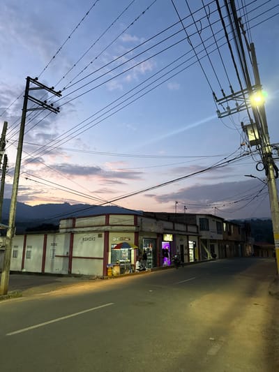 This image depicts a street scene in Timaná, Colombia, at dusk or early night. The sky above transitions from deep blue to lighter hues with subtle pink and orange clouds near the western horizon, where distant, dark mountains are visible. Numerous utility poles dominate the upper frame, supporting a dense network of overhead power lines and communication cables. A bright yellow street lamp is active on the utility pole on the right, illuminating the street below, and another less bright lamp is on the pole to the left.

The asphalt street, appearing damp in sections reflecting artificial light, slopes gently upwards towards the right. A faded white dashed line marks the center of the road. On the left, a single-story building with white walls and red trim features rectangular recessed panels. Adjacent to this, at a street corner, is a commercial building with a small vendor stall positioned on the sidewalk. This stall is sheltered by a colorful striped umbrella (red, yellow, blue, green), beneath which a seated individual, presumably the vendor, is present. Another person stands facing the stall, appearing to engage in an interaction. Two yellow gas cylinders are visible near the vendor stall.

Next to the vendor stall, a shop displays merchandise under vibrant purple neon lighting. A dark-colored motorcycle is parked directly in front of this shop, oriented towards the left side of the street. Further down the street to the right, additional multi-story commercial buildings line the road, some with illuminated windows or storefronts, contributing to the ambient light of the scene.