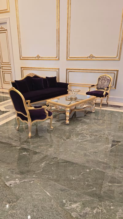 In the city of Agios Epiktitos, Northern Cyprus, an opulent living room features a deep purple velvet sofa with matching cushions and two ornate chairs, one on the left and one on the right. A gilded coffee table with a mirrored top sits in the center, adorned with a crystal centerpiece. The room is decorated with elaborate gold trim on the walls, and the floor is covered in polished green marble tiles. The overall ambiance suggests a luxurious and elegant setting.