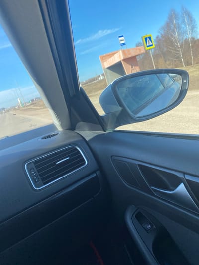 The image is taken from the interior of a vehicle, showing a side mirror reflecting a street scene. Visible in the mirror's reflection are road signs, buildings, and trees under a blue sky. The interior of the car features a window control, a vent, and the dashboard. The location is Verkhniye Tatyshly, Russia, as indicated by the bus stop sign written in Russian.
