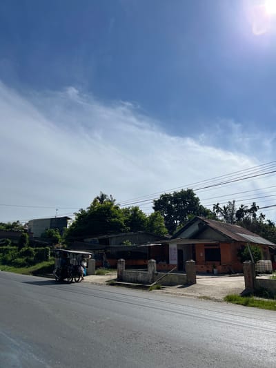 In Bukitrata, Indonesia, a street scene unfolds under a bright, partly cloudy sky. A paved road occupies the foreground, with the dark asphalt showing signs of wear. To the right of the road, a low-lying building with an orange facade and a rusted, corrugated metal roof is visible. The building has an open entrance and appears to be a small business or residence. A concrete walkway and a low wall with pillars lead up to the structure. In front of the building, a few potted plants add a touch of greenery. A group of people are gathered near the building, some standing by a small, covered vehicle that resembles a tricycle or auto-rickshaw, which is parked on the roadside. The vehicle has a canopy and appears to be carrying passengers. Behind the orange building and to the left, other structures and lush green vegetation, including palm trees, are present. Power lines are strung across the scene, adding to the urban or semi-urban environment. The overall impression is one of a typical day in a tropical locale, with everyday life continuing along a well-trafficked road.