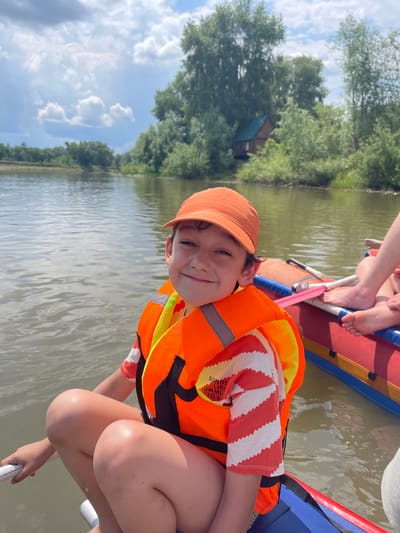 A young boy wearing an orange baseball cap and an orange life vest smiles at the camera while kneeling on a raft. He has on a white and orange striped shirt. The raft is on a body of water surrounded by lush green trees. In the background, a small wooden house with a blue roof can be seen among the trees. The sky is blue with fluffy white clouds. The image appears to be taken on a sunny day. The raft has other people on it, whose legs and feet are visible. The setting is likely a natural environment, possibly a river or lake, in Нежинка, Russia. The boy's expression suggests happiness and excitement. The life vest has reflective stripes. Partially visible text on the boy's shirt reads "SOUNDS LIKE SUMMER TO M".