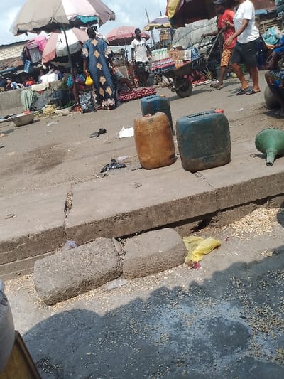 An open marketplace scene features vendors under umbrellas selling produce. Plastic containers sit atop a concrete ledge. In the foreground, textured concrete blocks lie on the ground amidst scattered debris, including a yellow plastic bag and fragments of organic material. The scene is lit by natural daylight. Pedestrians and vendors intermingle in the background.