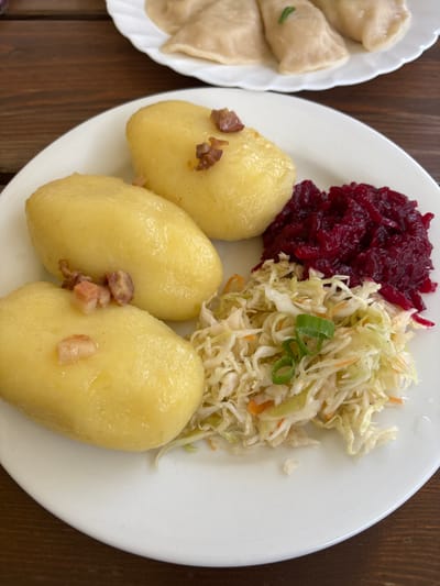 The image displays a close-up, elevated view of two plates of traditional Polish cuisine set on a brown wooden table, likely in Wojnowo, Poland. In the foreground, a white, round plate contains three large, elongated, light-yellow potato dumplings, each topped with small pieces of fried bacon or pork belly. Adjacent to the dumplings are two distinct piles of side dishes: a heap of dark red, shredded beetroot salad and a lower pile of light green shredded cabbage salad, garnished with slivers of orange carrot and chopped green onions. In the blurred background, a second white, round plate is partially visible, holding multiple light-colored pierogi, some adorned with small green herb garnishes. No individuals are present or visible in the scene.