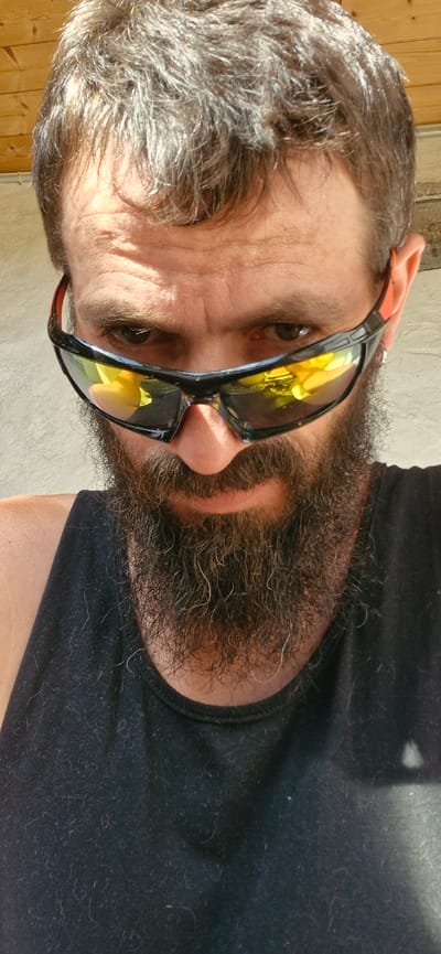A close-up, outdoor, selfie-style photograph shows a Caucasian man with a full beard and brown hair, wearing sunglasses and a black tank top. The man's face is tilted slightly downward and to his left, with his eyes looking directly at the camera through his sunglasses. The lenses of the sunglasses are reflective, showing yellow and orange hues, possibly from the surrounding environment or lighting. A faint silver hoop earring is visible in his right ear. The man's beard is thick and appears to be well-maintained. The background consists of a textured, light-colored wall and a wooden ceiling or structure above, suggesting an outdoor setting under a roof or awning. The lighting indicates it is daytime, with shadows cast across the man's face and the background. The image is likely taken in Wattenwil, Switzerland, given the context provided.