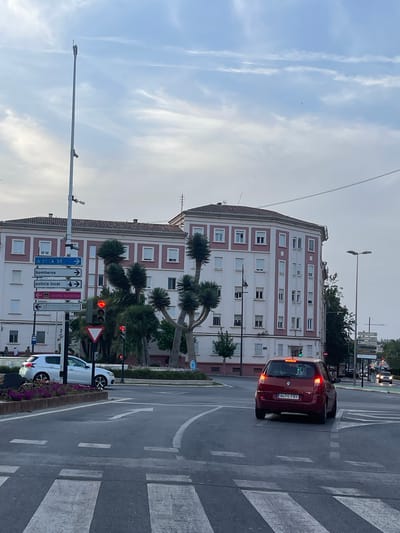 This image captures a street scene in Barriada San Ginés, Spain, during the day. The foreground shows a road intersection with a pedestrian crossing. A red car with license plate "8470 FRY" is stopped at a red traffic light. To the left, a white car is parked. In the background, a large, multi-story building with light pink and white facade stands. A tall pole with various directional signs is visible on the left side of the road, including signs for "A-37 A-30", "bomberos", "policia local", and "centro histórico". Palm trees and other greenery are planted around the intersection. The sky is partly cloudy.