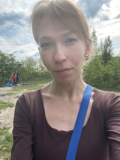An outdoor selfie captures a young woman with light-colored hair and blue-green eyes, looking directly at the camera with a neutral expression. She is wearing a dark brown long-sleeved top, and a bright blue strap, possibly from a bag, crosses her body. A gold-colored earring is visible in her right ear.

The background shows a natural outdoor setting under a cloudy but bright sky. Prominent green trees and bushes frame the scene, with the ground composed of dirt, gravel, and patches of grass. In the middle distance, three other individuals are visible. Two appear to be standing, one dressed in dark clothing with red accents, and the other predominantly in black. A third person is partially obscured, possibly kneeling or sitting on the ground next to a blue object.

The scene suggests a daytime outdoor activity, such as a casual walk or gathering in a natural, semi-rural environment. The weather is bright with clouds. The location is in an outdoor area, specifically "Пятый километр, Russia". No discernible text is visible in the image.
