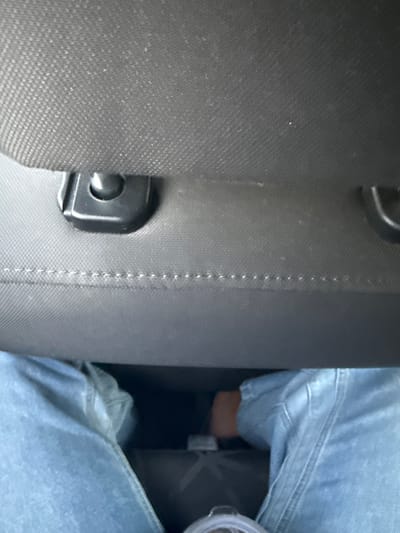 The image depicts the interior of a passenger vehicle, observed from a low angle, likely from a rear passenger seat looking forward. The upper two-thirds of the frame are dominated by the rear of a dark grey or black fabric car seat, including its headrest. Two dark, likely plastic, headrest post anchors are visible near the top. A distinct horizontal line of white stitching runs across the seatback fabric, marking a seam.
In the lower third of the frame, the lower extremities of an individual are visible, wearing light blue denim jeans. The legs are slightly parted, revealing a dark void towards the vehicle's floorboard. Partially visible on the floorboard, between the legs, is a dark object, possibly a bag or mat, featuring a subtle lighter-colored "X" pattern. In the bottom-center foreground, a clear plastic cup with a translucent lid is partially visible. To the right, within the dark recess near the right leg, a patch of light brown material, consistent with skin or light-colored footwear, is discernible.
The scene is confined to the vehicle interior. The image contains no external geographical markers, landmarks, or identifying features that would allow for the precise determination of its location within the city of West Windsor, United States, or any other specific geographic area. The evident subject is a person seated as a passenger in a vehicle. No specific actions or interactions are discernible beyond being seated.