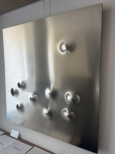An art installation is documented within an interior space located in Galatina, Italy. The image features a large, vertically oriented, rectangular panel of brushed or semi-polished metallic material, affixed to a plain, light-colored wall. The panel exhibits approximately ten distinct, irregularly spaced conical indentations or protrusions across its surface. Each such feature is centered by a small, dark metallic pin, generating localized reflective light patterns. The panel is suspended from a ceiling-mounted track system by thin cables. Below the panel, on a light-toned surface, are multiple white, softcover booklets; one visible cover reads "levelProject." Adjacent to the booklets, a small, clear acrylic display stand holds text, which is not legible. The ceiling above the artwork is a light brown, possibly wood-grained material.