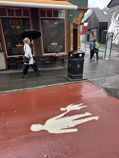This image captures an outdoor street scene in downtown Reykjavik, Iceland, on a rainy, overcast day. The perspective is slightly elevated, looking down a street with buildings on either side.
On the left, a two-story building with a green corrugated metal facade and an orange awning dominates. Its ground floor features two large windows for the "ELLY" bar, displaying "BEER FOR A GOOD TIME" and "LAVAZZA" branding in gold script. Chalkboard-style menus below the windows list items like "KARAOKE," "HAPPY HOUR," "COCKTAILS," and "BAR FOOD" in Icelandic. Black metal chairs and small tables are arranged on the wet grey pavement in front of the bar. A blonde-haired woman, wearing a white tunic and dark pants, holds a black umbrella overhead while walking from right to left on this pavement. A second person, largely obscured, walks behind her in the same direction.
In the mid-ground, a black public trash can, labeled "BLANDAÐUR ÚRGANGUR" (Mixed Waste) with recycling symbols, stands on the grey pavement. Behind it and to the right, a narrow street or alleyway leads uphill. Two individuals wearing hooded raincoats (one purple, one dark green with a light green backpack) are walking away from the viewer up this street. A green signpost in the mid-ground displays "SVÆÐI" (Area) and "GÖNGUGATA" (Pedestrian Street). Buildings with varying facades, including a red structure and a dark grey corrugated metal building with a skylight, line this street. On the far right, a large, abstract white mural or sculpture is visible on a stone wall.
The foreground is covered by a wet, reflective red-painted surface, likely a dedicated pedestrian or bicycle path. Two stylized white human figures are painted on this red surface, appearing to be lying down or "falling." The larger figure is in the bottom left, lying on its back with arms and legs splayed, while a smaller figure is positioned above and to its right, also on its back. The entire ground surface, including the grey pavement and the red path, is visibly wet with puddles and reflections, indicating recent or ongoing rain.