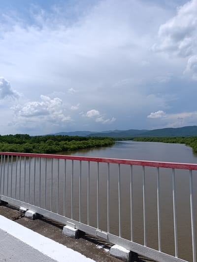 A wide-angle, eye-level shot captures a river flowing through a lush green landscape under a cloudy sky. The foreground is dominated by a white metal railing with a red top, part of a bridge. The railing extends across the bottom and middle sections of the image. Beyond the railing, the calm, brown river stretches into the distance, bordered by dense green trees that form a thick, continuous line along the banks. In the far background, rolling hills covered in trees recede into the hazy horizon. The sky is a mix of bright blue and large, fluffy white clouds, with some darker, more ominous clouds present. The image appears to be taken from a bridge in or near the city of Kazanka, Russia. There are no people or animals visible in the frame.
