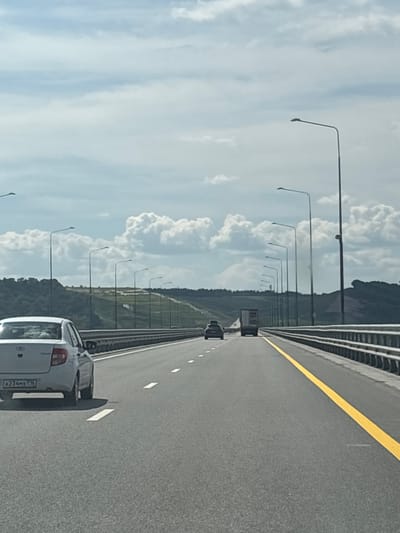 The image depicts a view from a moving vehicle on a highway, likely outside the city of Orel, Russia, based on the license plate of the car in the foreground. The road surface is asphalt with clear white lane markings and a bright yellow line on the right side. A white sedan with a license plate reading "A234MX 716" is visible in the left lane in the foreground, its rear end and side facing the camera. Further ahead in the same lane, another car is visible. In the right lane, a truck is moving away from the camera, followed by other vehicles. The highway is flanked by a metal guardrail and a row of lampposts stretching into the distance. The background features a hilly, green landscape with trees and a partly cloudy sky. The overall scene suggests a typical daytime commute on a major road.