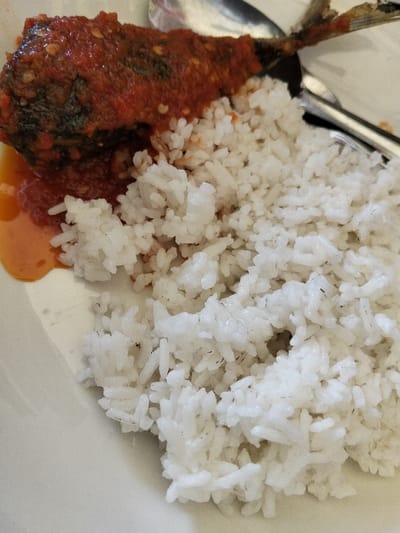 The image presents a close-up view of a meal served on a plain white plate, originating from Keudeteunom, Indonesia. The main components of the dish are a large portion of white rice and a piece of fish generously coated in a vibrant red chili sauce, known as sambal.
The fish, which appears to have been fried, is almost entirely covered by the thick, glossy red sambal. Hints of its darker, possibly crispy skin are visible underneath the sauce, which features visible chili flakes and textural elements. A small pool of orange-red liquid, likely sauce or spiced oil, has seeped from the fish and collected on the white plate in the bottom-left corner.
Adjacent to the sauced fish is a mound of fluffy, white, cooked rice. The individual grains are distinct, suggesting it is well-cooked and fresh. The rice appears slightly moist and glistening in parts. A metallic spoon is partially visible in the upper right quadrant of the image, resting on the plate, indicating the meal is ready for consumption.
The lighting is bright and even, illuminating the food clearly and enhancing the vibrant colors of the sambal and the whiteness of the rice. No people are visible in the frame, nor is there any discernible text. The dish is characteristic of Indonesian cuisine, commonly featuring fish prepared with spicy sambal and served with rice.