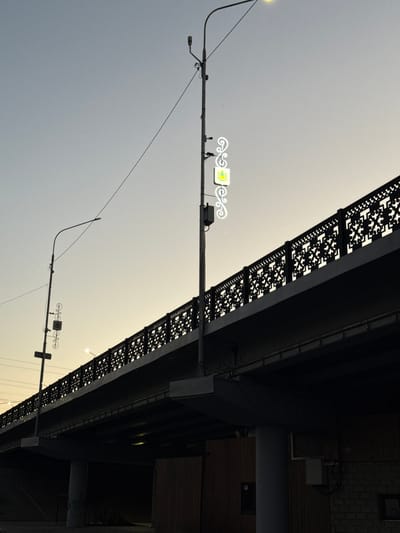 A low-angle, wide shot captures a bridge spanning the sky in the city of Kamysh-Gadzhiaul, Russia, at dusk. The sky is a pale gradient of blue and yellow, transitioning to a darker blue overhead. A tall, slender streetlamp stands to the left of center, its curved arm holding a light fixture at the top and adorned with decorative white neon elements and a small, square emblem. The bridge, a dark silhouette against the sky, features an ornate black metal railing with a repeating geometric pattern. Beneath the bridge, the scene is largely shadowed, revealing structural elements and wooden-clad walls of a building. The overall impression is one of urban infrastructure set against a tranquil, fading light.